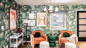 Ein Wohnzimmer mit floralem Wandpapier, zwei orangefarbenen Sesseln, einem grünen Beistelltisch und einer weißen Regal, auf der Bücher und eine rote Tasse stehen.