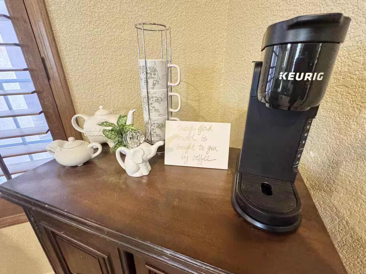 A coffee maker, teapot, and cups on a wooden table with a card.