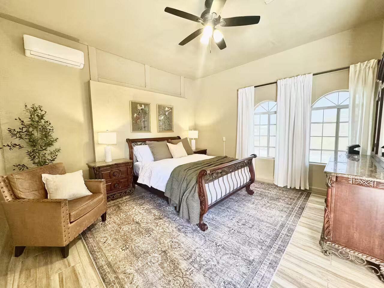 Une chambre à coucher comprenant un lit, une commode en bois, un canapé, un ventilateur de plafond et une grande fenêtre avec des rideaux.