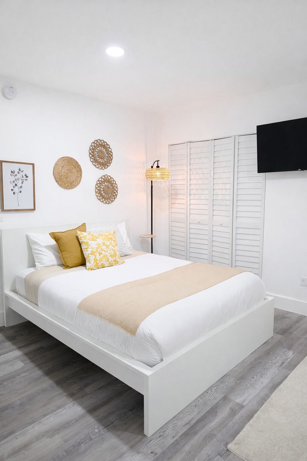 A modern bedroom with a white bed, a bedside lamp, a framed artwork, and a television on the wall.