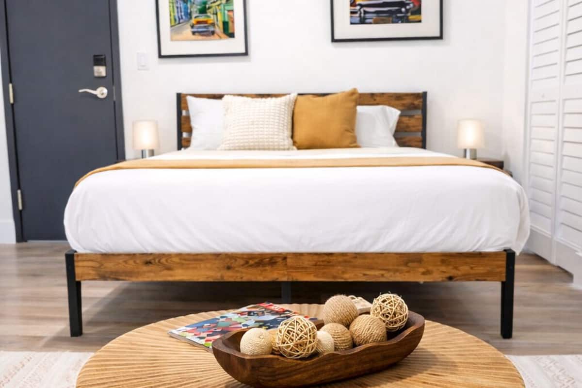 A modern hotel room with a wooden bed, two framed pictures on the wall, a round wooden coffee table with decorative balls, and a patterned rug.