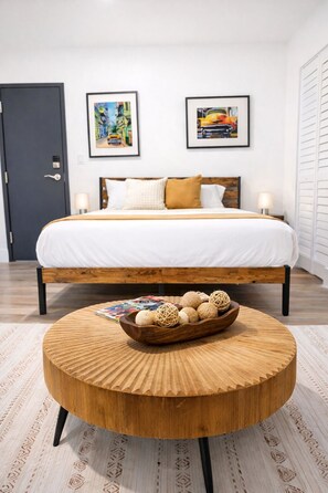 Une chambre d’hôtel moderne avec un lit en bois, deux tableaux encadrés au mur, une table basse ronde en bois ornée de boules décoratives et un tapis à motifs.