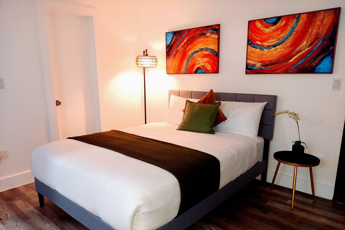 A bed with a white and brown bedspread, two pillows, and a headboard. There are two paintings on the wall and a floor lamp.