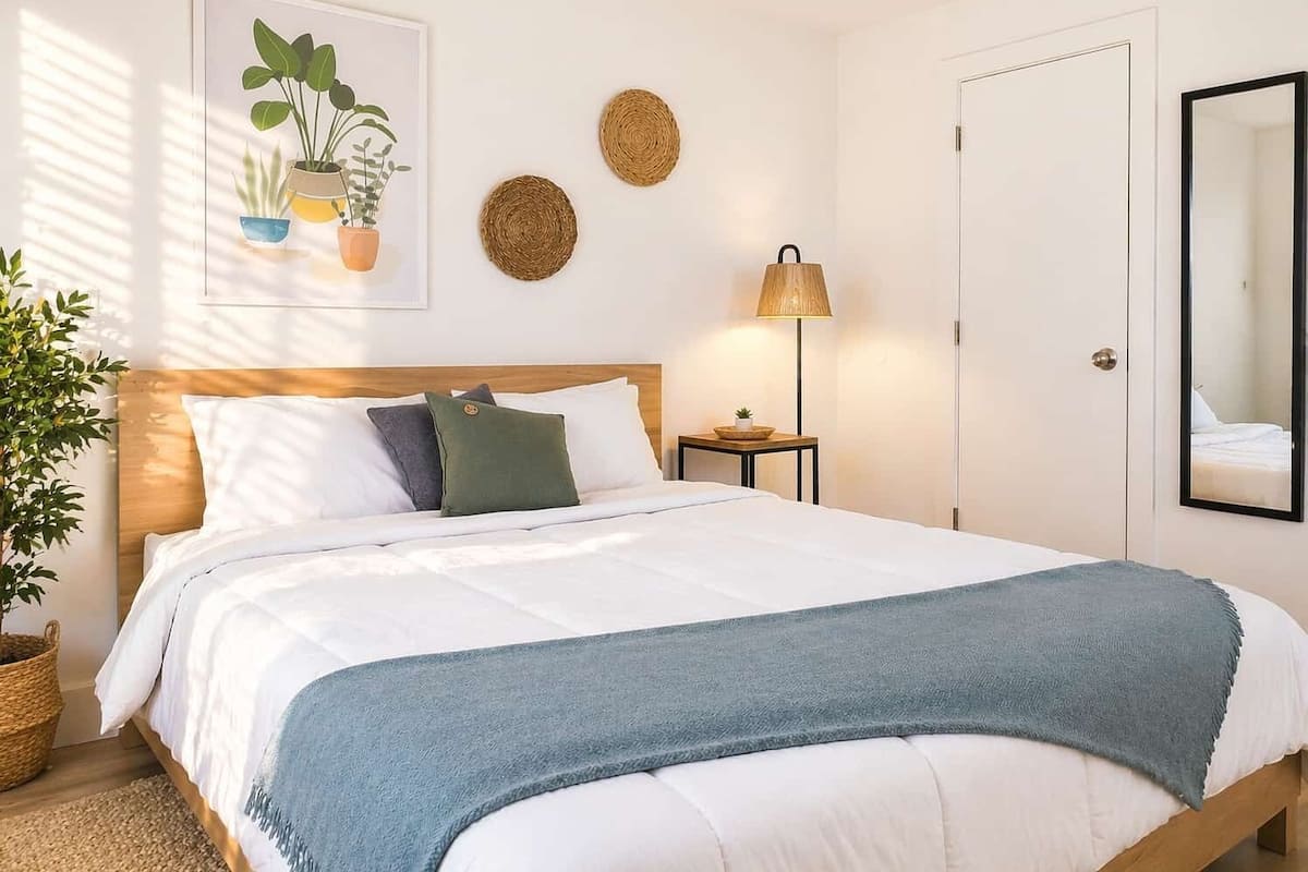A neatly made bed with a blue blanket, a bedside lamp, and a framed plant on the wall.