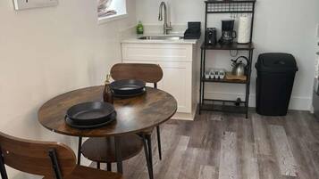 A compact kitchen with white cabinets, a dining table with chairs, a coffee machine, and a small shelf with kitchen items.