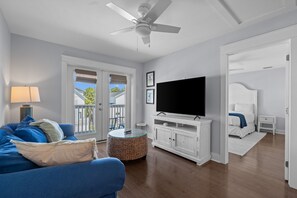 Ein Wohnzimmer mit blauem Sofa, einem Fernseher auf einem weißen Schrank, einem Deckenventilator und Blick ins Freie durch eine Tür.