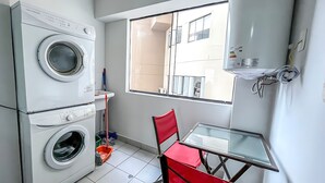 Une petite buanderie avec des lave-linge et des sèche-linge superposés, une petite table avec une chaise et une fenêtre donnant sur des immeubles.