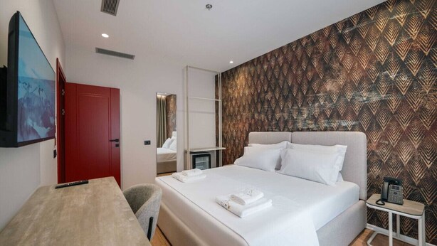 A modern hotel room with a large bed, a TV mounted on the wall, a red door, and a patterned wallpaper. - Ermes Deluxe Family Room With French Balcony (Durrës)