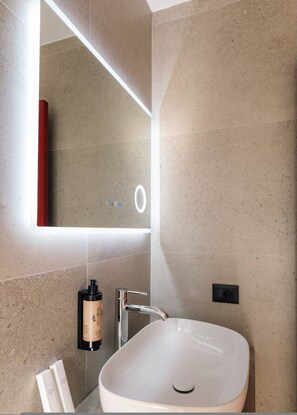 Ein modernes Badezimmer mit einem großen Spiegel, einem weißen Waschbecken und einer Shampooflasche.