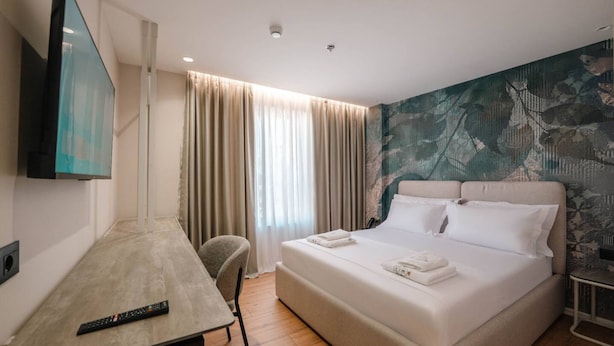 Ein Hotelzimmer mit Bett, Schreibtisch, Stuhl, Fernseher und einem großen Fenster mit Vorhängen.