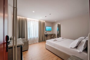 A hotel room with a bed, a desk, a chair, a TV, and a large window with curtains. - Ermes Deluxe Double Room With Sofa Bed (Durrës)