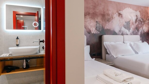 Ett modernt hotellrum med en stor, vit tvättställ, en spegel med inbyggd belysning, en säng med vita sängkläder och en abstrakt väggmålning med en intressant struktur.