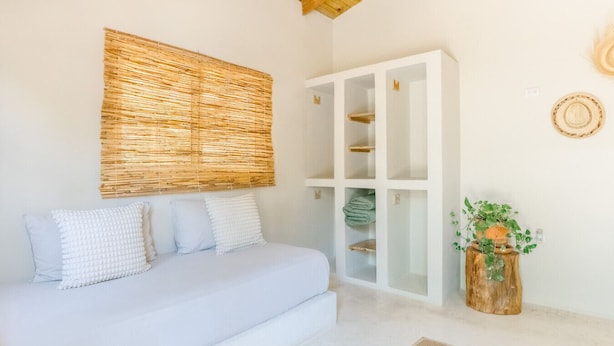 Una camera da letto con un letto, un armadio bianco, una testiera in fibra naturale e un piccolo sgabello di legno con una pianta.