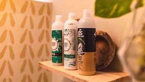 Tre bottiglie di Shampoo Coco Biotin su un ripiano di legno, con una parete decorata con un motivo a foglia.