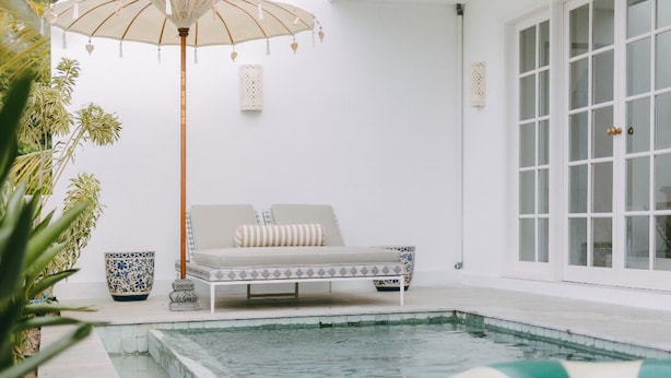 Area piscina con una poltrona da relax e un ombrellone decorativo, circondata da vegetazione e un edificio moderno con ampie finestre.