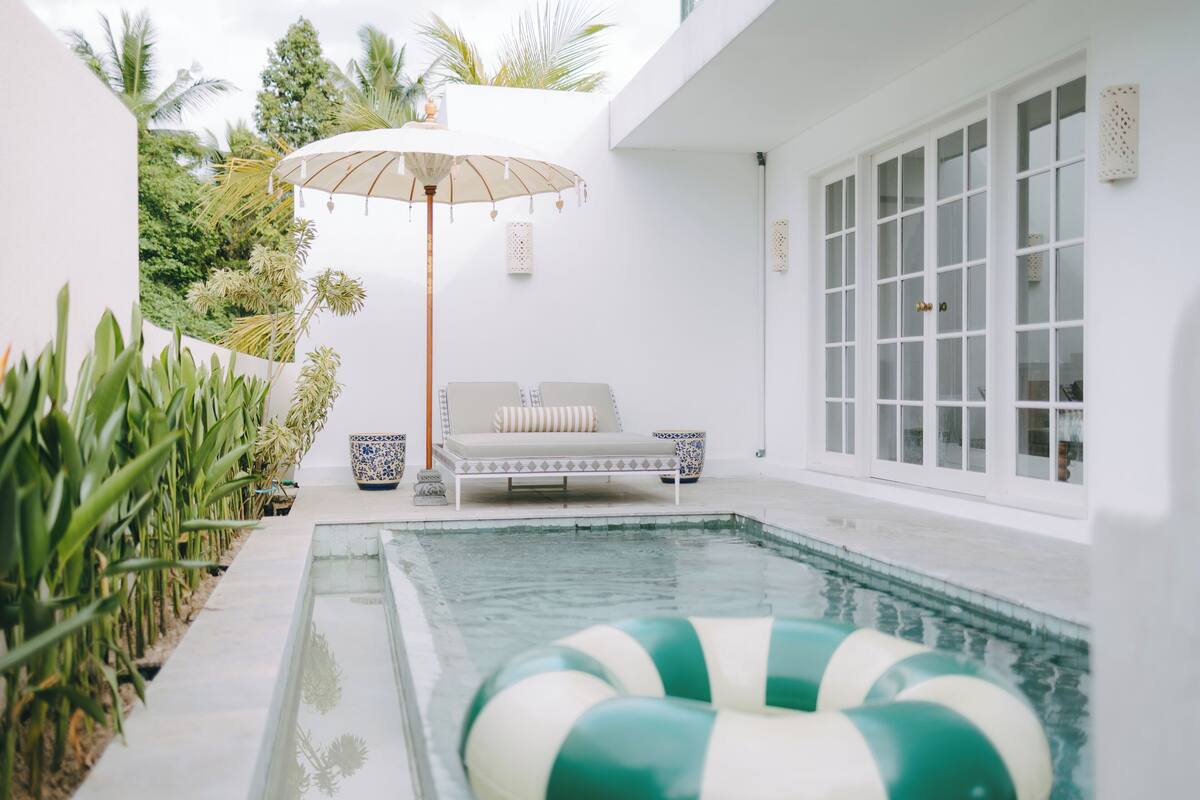 A modern outdoor pool area with a lounge chair and umbrella, surrounded by greenery.
