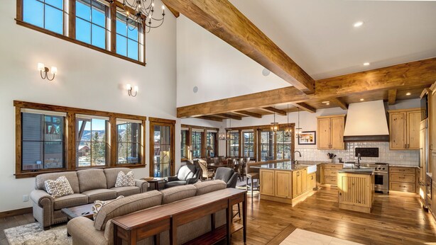 A spacious living room with a large sofa and wooden coffee table, a kitchen with wooden cabinets and a central island, and large windows offering views of the outdoors. - Brand new, 5BR home with private shuttle and concierge (Steamboat Springs)