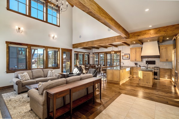 A spacious living room with a large sofa and wooden coffee table, a kitchen with wooden cabinets and a central island, and large windows offering views of the outdoors. - Brand new, 5BR home with private shuttle and concierge (Steamboat Springs)