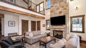 A spacious living room with a fireplace, a large flat-screen TV, and a high ceiling with exposed beams. - Brand New! 8BR home, private shuttle and concierge (Steamboat Springs)