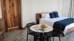 Hotellihuone, jossa on sänky, pöytä ja tuolit sekä puinen ovi ja ilmastointilaite.