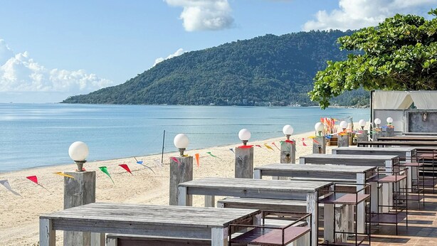 Un restaurant situé en bord de mer, avec une terrasse, une vue sur l’océan et une colline boisée en arrière-plan.