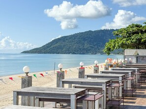 A beachside restaurant with outdoor seating, a view of the sea, and a forested hill in the background.