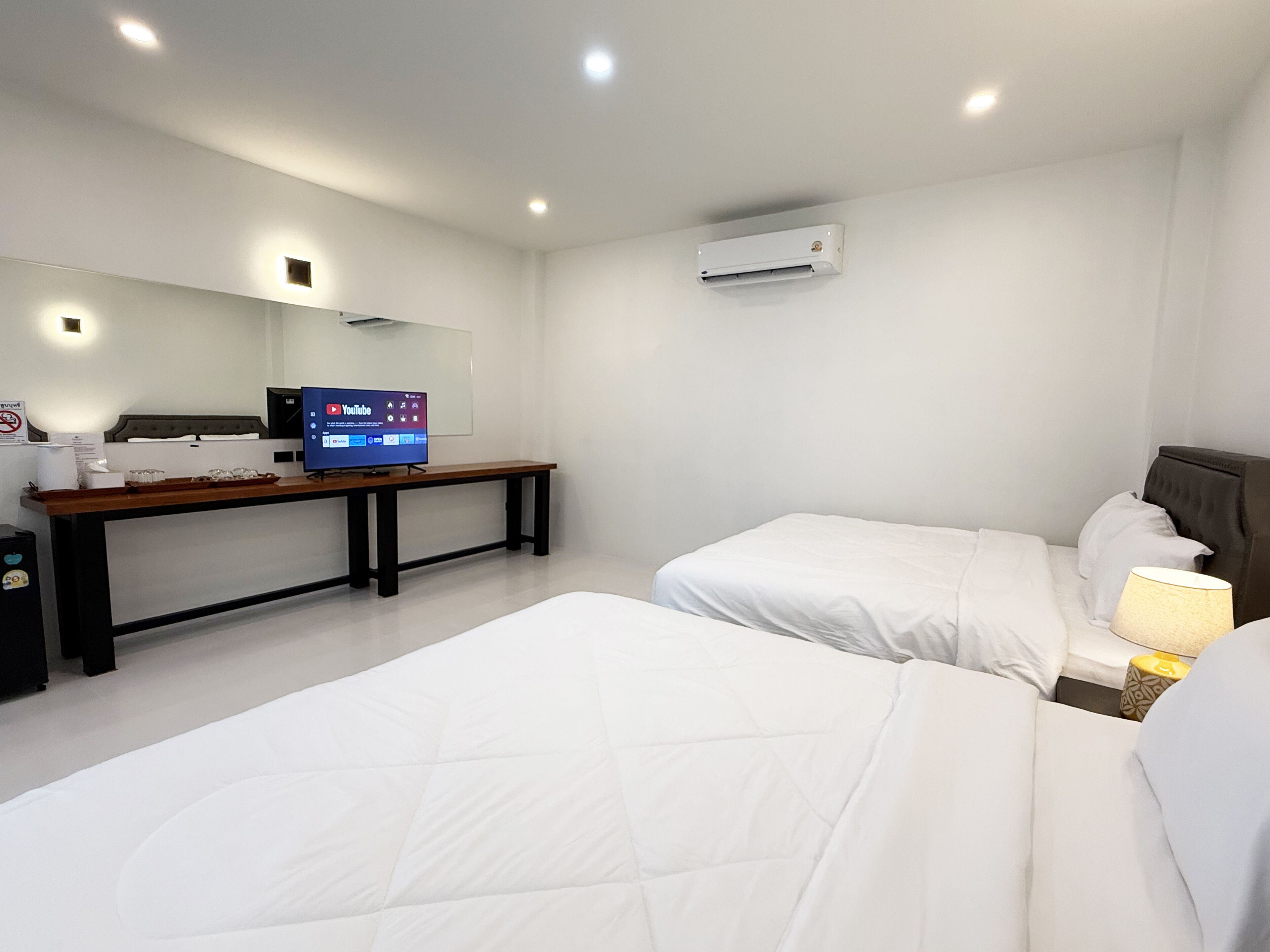 A hotel room with two beds, a desk with a computer, a TV displaying YouTube, and an air conditioner on the wall.