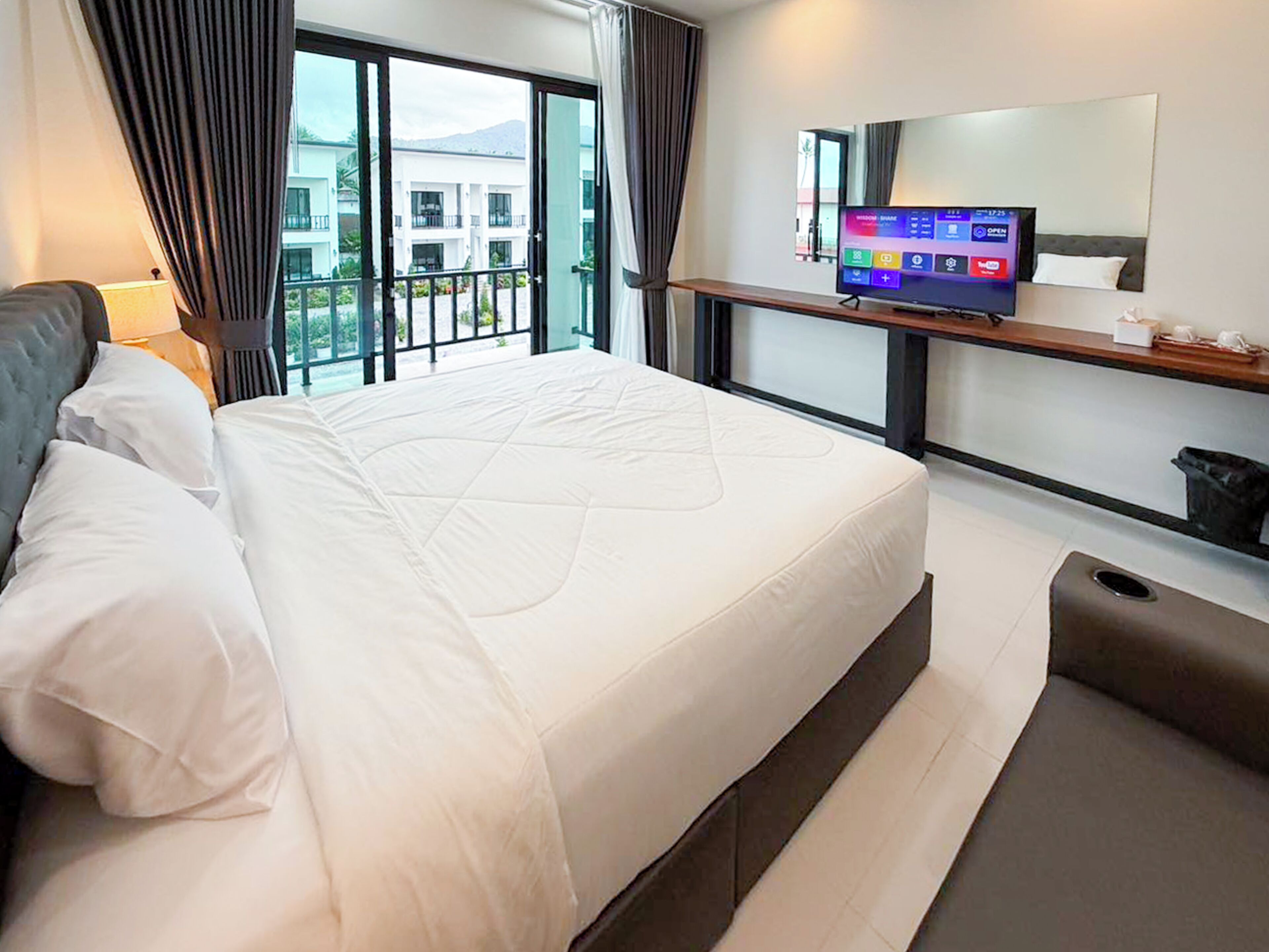 Habitación de hotel con una cama grande, un televisor sobre un mueble, una ventana con cortinas y vistas a edificios.