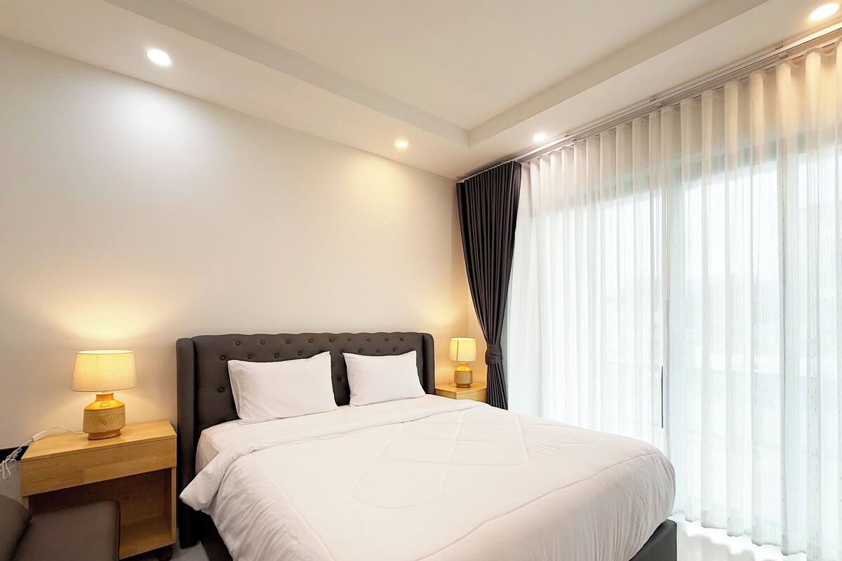 A modern hotel room with a large bed, two bedside tables with lamps, and a large window with sheer curtains.