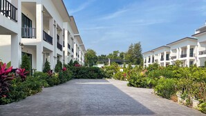 Une cour avec un chemin pavé, entourée d’une végétation luxuriante et de bâtiments blancs dotés de balcons.