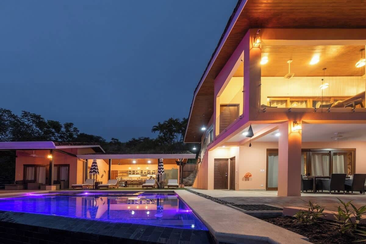 A modern two-story house with a swimming pool, outdoor seating, and a well-lit interior.