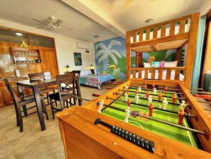 Ein Zimmer mit Fußballtisch, Holzmöbeln, einem Palmen-Gemälde und Deckenventilator.