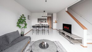 A modern living room with a grey sofa, a coffee table, and a TV displaying 'NETFLIX'. There's a dining area with a table and chairs in the background.