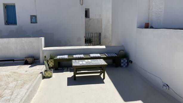 Un pequeño espacio al aire libre con mesa y bancos, rodeado de paredes blancas y un portón de metal.