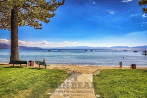 Ein Strand mit einer Bank, einem Baum und freiem Blick auf das Wasser und die dahinter liegenden Berge.