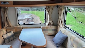 A cozy RV interior with a central table, cushioned seating, and a large window offering a view of a grassy area.
