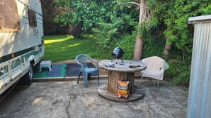 A campsite with a trailer, a round wooden table with charcoal, two plastic chairs, and a small stool.