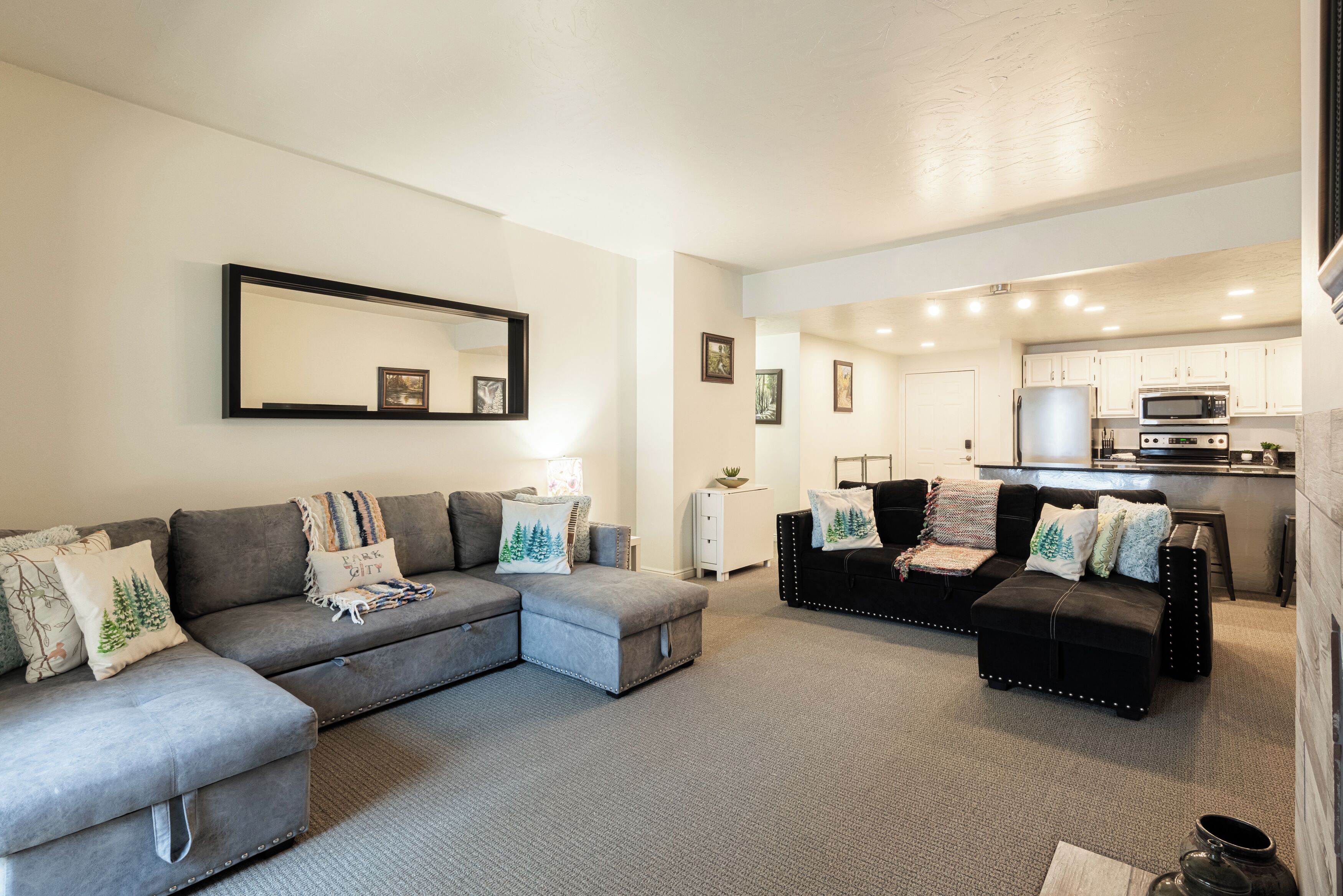 A modern living room with a grey sectional sofa, a black leather sofa, and a white side table. A kitchen is visible in the background with white cabinets and stainless steel appliances.