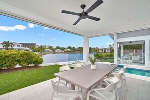 Eine moderne Außenpatio mit Esstisch, weißen Stühlen, Deckenventilator und Blick auf einen See sowie umliegende Gebäude.