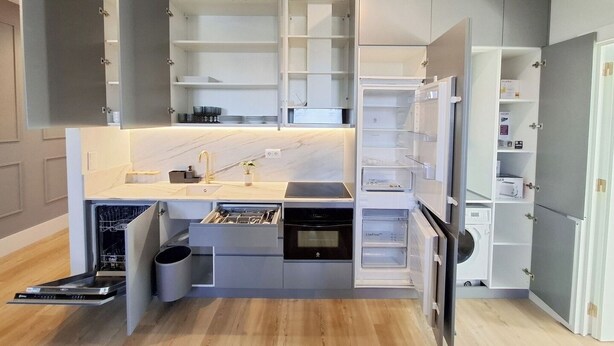 Une cuisine compacte dotée d’appareils électroménagers intégrés, notamment un lave-vaisselle, un four et un réfrigérateur, ainsi que d’étagères et de placards ouverts pour le rangement.