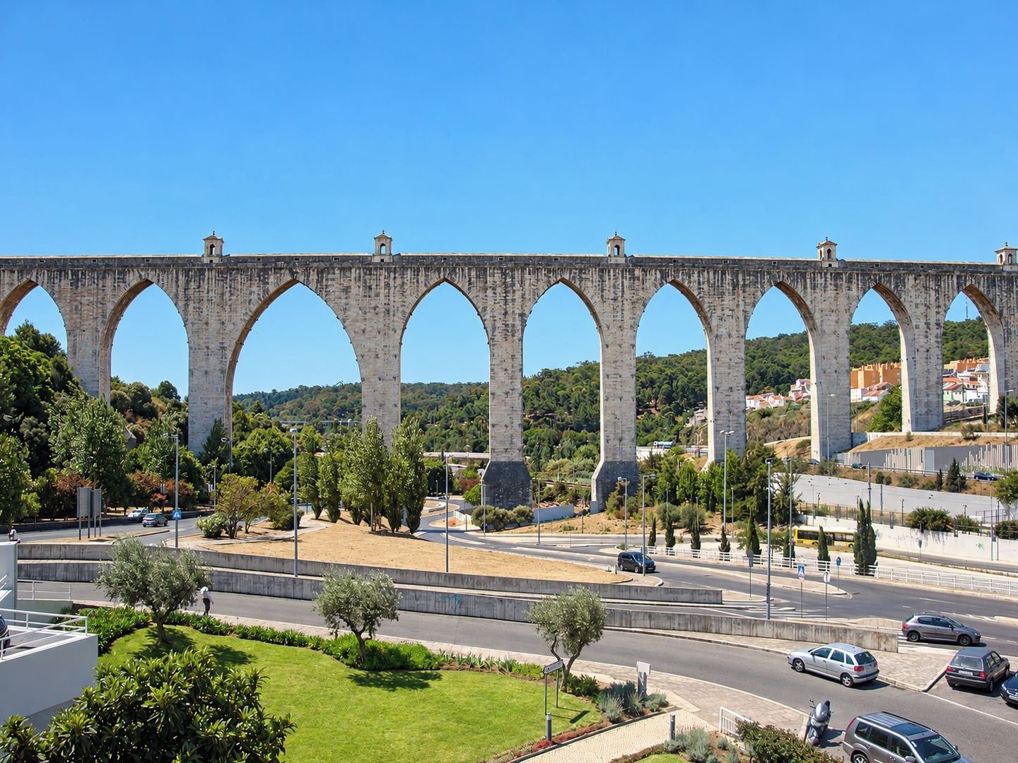 Uma ponte de pedra com várias abóbadas sobre uma estrada, rodeada por vegetação e edifícios residenciais.