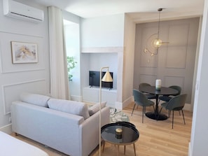 Ein modernes Wohnzimmer mit grauem Sofa, einem Esstisch mit vier Stühlen, einem Fernseher und einer Pendelleuchte.