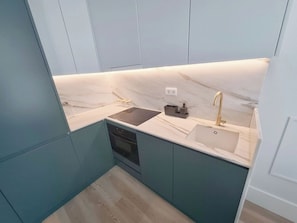 Eine kleine Küche mit türkis-farbenen Schränken, einer schwarzen Induktionskochplatte, einem Waschbecken mit goldfarbenem Wasserhahn und einer Marmor-Spritzschutzwand.