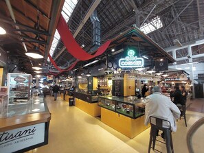 Ein überdachter Markt mit verschiedenen Ständen, darunter ein Schild mit der Aufschrift 'Gin Corner' und ein Stand namens 'Japonés'.