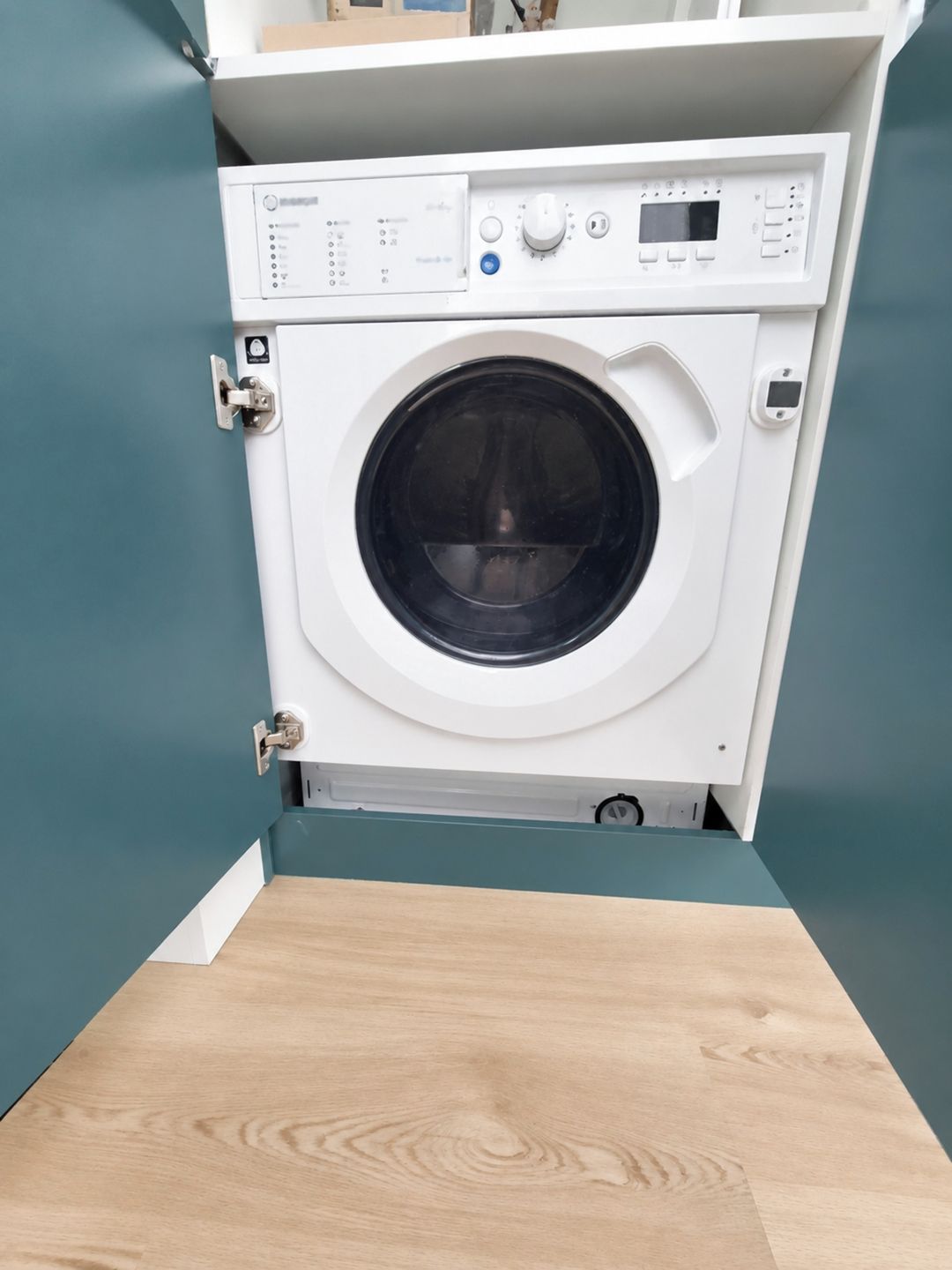 A compact, white front-loading washing machine in a corner with teal walls and a wooden floor.