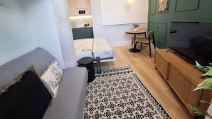 Un espacio habitable compacto con sofá, una pequeña mesa de comedor con sillas, televisor y una kitchenette al fondo.