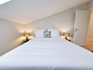 Ein Zimmer mit einem Einzelbett, geneigter Decke, zwei Nachttischlampen, einer Tür rechts und einem gemusterten Kopfkissen auf dem Bett.