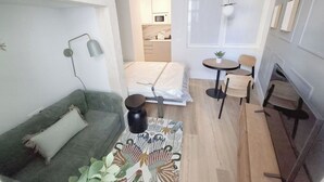 Een ruimtelijke inrichting met een groene bank, een bed, een kleine ronde tafel met stoelen, en een keukenhoek zichtbaar door een open deur.