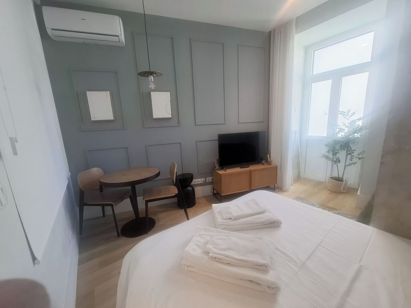 Une chambre d’hôtel avec un lit, une télévision, une petite table avec une chaise, une plante en pot et une fenêtre.