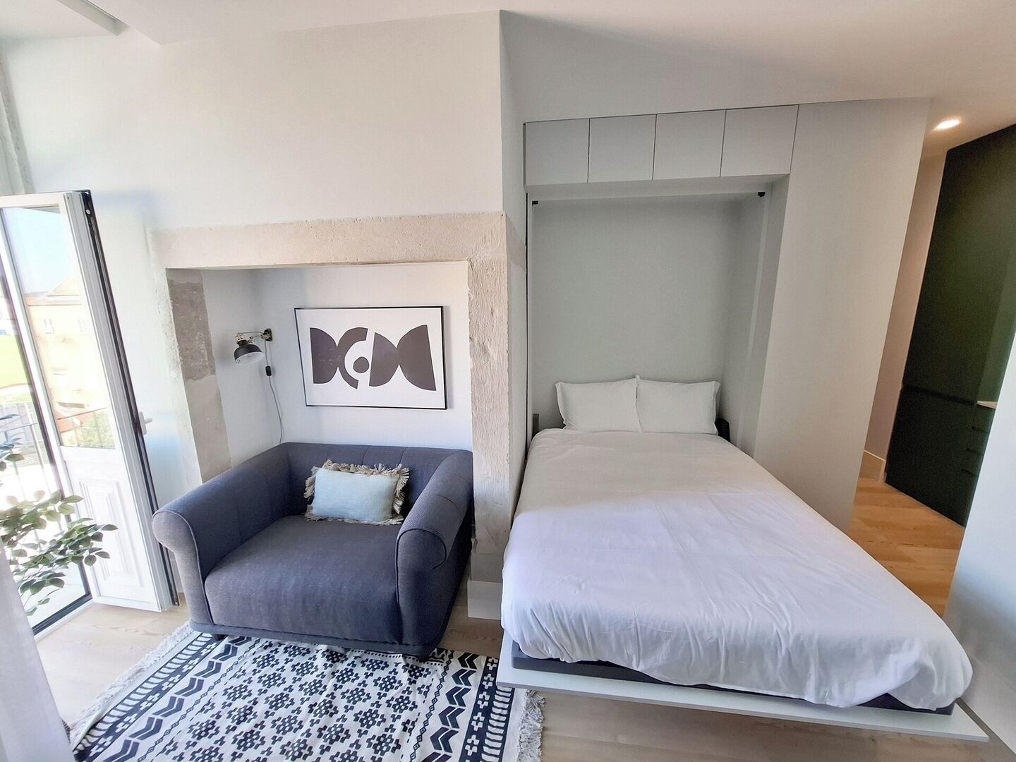A modern bedroom with a bed, a gray armchair, a patterned rug, and a wall-mounted artwork.